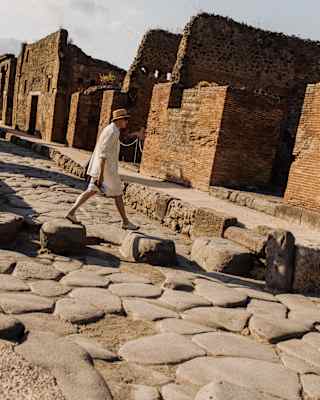 A man in a straw hat and loose white clothing strides across an ancient cobbled street lined with dramatic ruins at Pompeii.