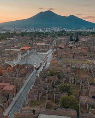Aerial view of the ancient ruins of Pompeii with Mount Vesuvius in the background at sunset, showcasing stone streets and remains of buildings.