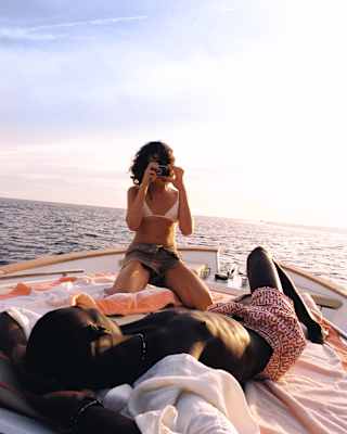 A woman raised up on her knees photographs her lounging companion as they relax on towels on the deck of a private boat.