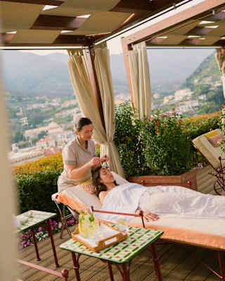 A spa therapist sits at the head of a treatment bed where she massages the forehead of a guest who lies beneath a sheet.