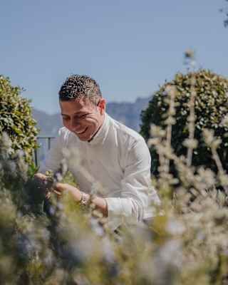 Chef Armando Aristarco wears long sleeve chef whites and smiles as he picks fresh oregano from the hotel gardens