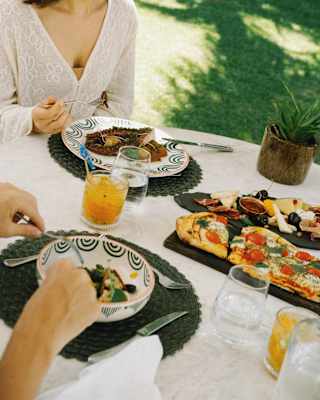 Angled close-up of two guests enjoying light lunch at an al-fresco garden table with pizza, grilled octopus and cheese board.