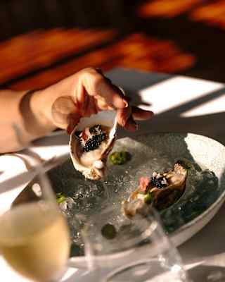 A guest raises a half-shell oyster from a bowl of ice, seen close-up at a table with white wine and water, at Restaurant Y.