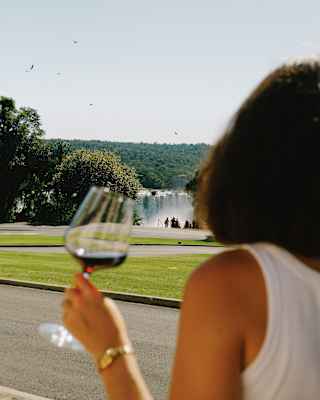 A group people stand at a waterfall viewpoint on the other side of lawns, seen from behind a woman with a glass of red wine.