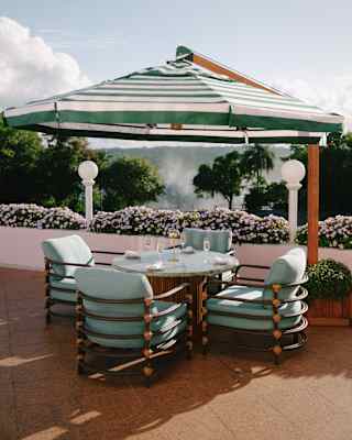 A large green-striped cantilever parasol shades a round table with four wine flutes and pale blue chairs overlooking gardens.