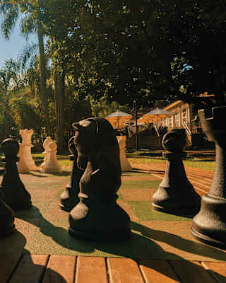 Large black and white chess pieces are set up on a giant outdoor chessboard, surrounded by trees and sunlight, with a building visible in the background.