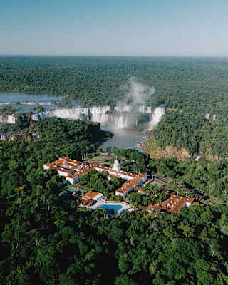 Aerial view of a resort surrounded by dense forest, with large waterfalls and mist rising in the background under a clear sky.