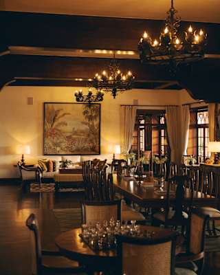 Three candle chandeliers glow above a polished table with chairs and flower vases, seen over a small table with a chess set.