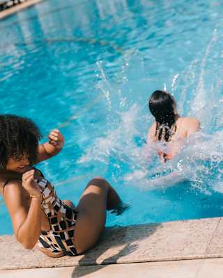 A dark-haired woman on the pool edge shields herself as a friend in the water makes a splash, taken from behind on an angle.