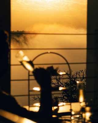 A hand raises a wine glass at a table with a cake stand in soft-focus, with a backdrop of gold light and waterfall spray.
