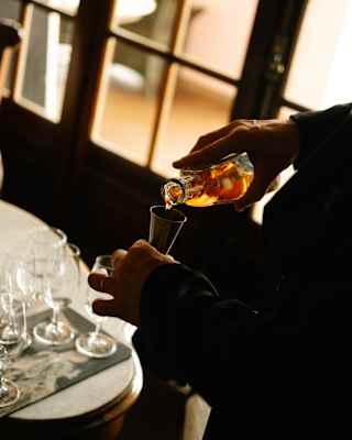 A staff member pours amber Cachaça from a bottle into a measuring cylinder above a table of tasting glasses, in a close-up.