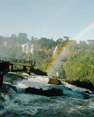 Angled image of a woman facing high cascades as she stands on a viewing stage over a river with a rainbow arc in its spray.