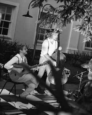 Two musicians perform outdoors; one sits playing guitar while the other stands playing a double bass. Both wear light shirts and braces. Sunlight and shadow fall across the scene. A building with windows is visible in the background.