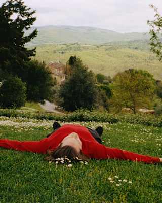A man in a red jumper lies like a starfish on a hill, feet pointing to the wide Tuscan views, seen from grass above his head.