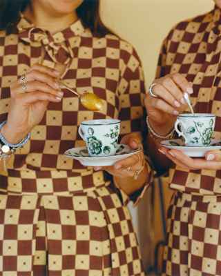 Two women in identical brown and white-checked dresses stir coffee in delicate cups, side by side, seen close-up.