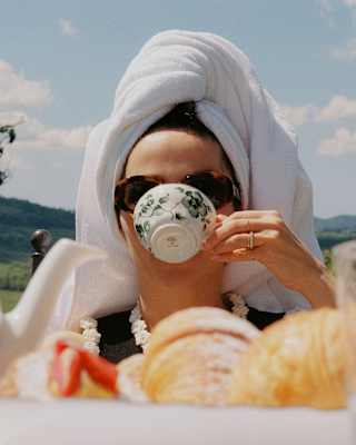 A woman with a towel hair-wrap drinks tea from a porcelain botanical-print cup, seen at eye-level over a table of pastries.