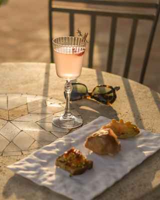 A plate of three savoury bites sits by a delicate pink drink which glows in the rays of late sun atop an al-fresco table.