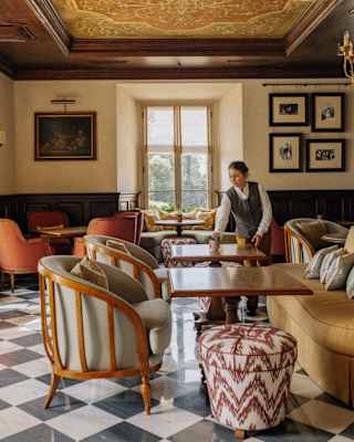 A waitress adjusts a table in the elegant Bar Visconti, with monochrome tiled floor, wood-framed tub chairs and sofas.