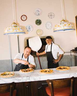 A chef garnishes one of four cooked pizzas on a marble table, while another holding a paddle stands in front of the oven.