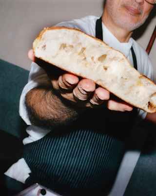A chef holding a baker's paddle presents a slice of bouncy focaccia-style bread, fresh from the oven, seen in close-up.