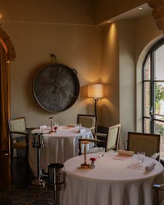Elegant restaurant interior with round tables covered in white cloths, set for dining. Soft lighting, a large decorative piece on the wall, wooden ceiling beams, and a window letting in natural light.