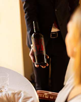A waiter, seen from the chest down, presents a bottle of red wine to a female diner in soft-focus, viewed over her shoulder.
