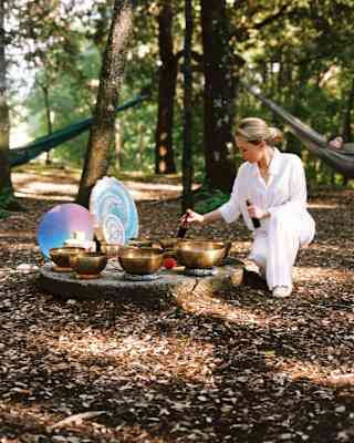 In a forest clearing, a woman in white taps metal singing bowls placed on a slab alongside other sound healing instruments.