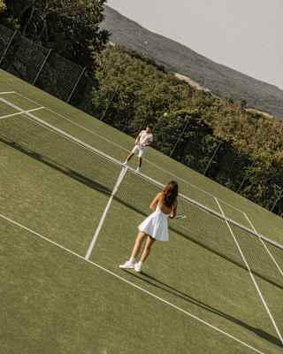 Angled image of a man and woman in tennis whites as they play on a grass court with forest views, seen from behind the woman.