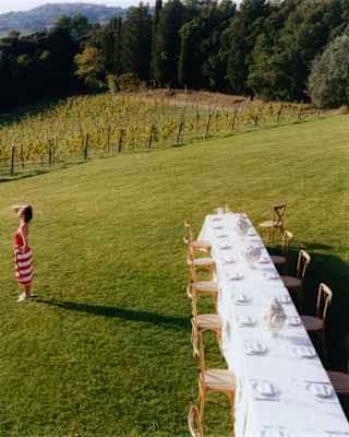 On the lawn, a woman in a red and white towel gazes at the view as she stands near a long celebration table, seen from above.