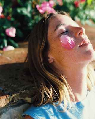 Close-up of a woman with blond hair and freckles as she leans back on a wall, face in sun, with a pink petal on one cheek.