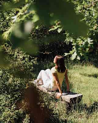 A woman leans back on her arms as she sits, feet up on a log bench in a grassy clearing, seen from behind through oak leaves.