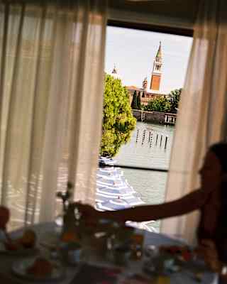 A couple inside a suite, in low light, enjoy breakfast by a window with crisp views over the canal to San Georgio Maggiore.