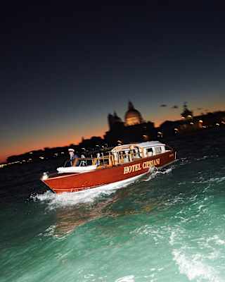 The Hotel Cipriani private launch cuts a bow wave through the green water at dusk. In the background, the lights of St Marks