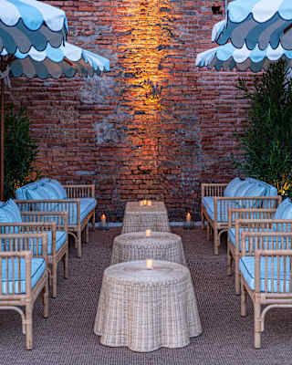 Two rows of three cane and blue chairs face each other over candle-lit rattan tables in a brick-walled bar area, seen at dusk.