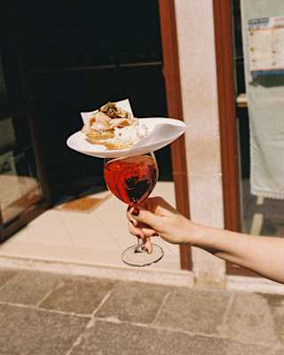 A plate of a cicchetti pastry treats rests on the top of a glass of iced Aperol, carried in a Venice street, seen close-up.