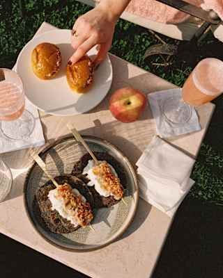 A woman's hand reaches for one of two paninis at a table with Peach Bellinis and savoury skewers on bread, seen from above.