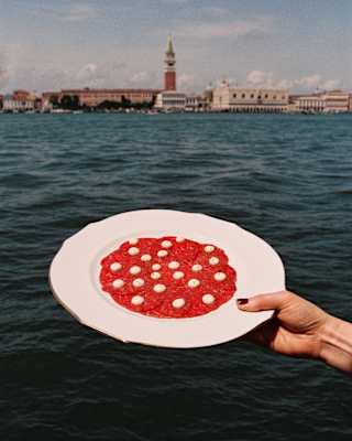 A woman's hand holds up a plate of Carpaccio dotted with Cip's sauce against the backdrop of the Canale della Giudecca.
