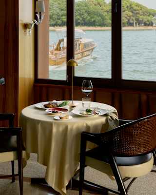A meal awaits guests at a pretty table for two next to windows overlooking the water, where a small wooden boat sails by.