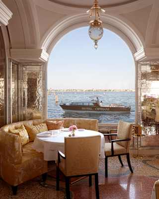Elegant dining table with gold chairs and sofa set by a large arched window, overlooking a calm body of water with a Hotel Cipriani boat passing by outside under a clear sky.
