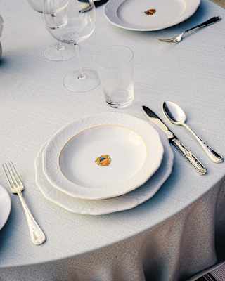 A round table set with a white tablecloth, stacked white plates, a fork, knife, and spoon, clear glassware, and a striped cushioned chair.
