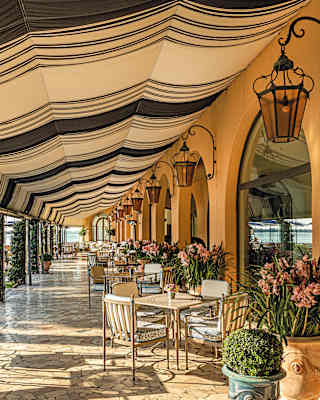 View along the tiled Caffè San Giorgio terrace where tables sit in dappled sun beneath a vast black and white canvas awning.