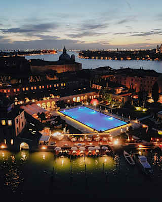 Aerial night view of Hotel Cipriani, sublit pool glowing blue and lights twinkling on Venice's docks, homes and landmarks.