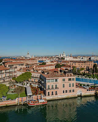 Aerial view of Hotel Cipriani's front, where a boat waits by striped mooring posts, and glimpses of greater Venice beyond.