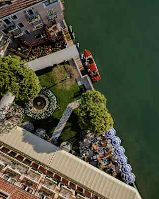 Birds-eye view of the hotel's white canopied pathway entrance, crossing lawn to the mooring posts, where a red boat waits.