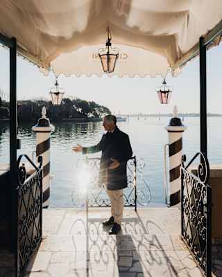 Sun dazzles on the water in front of the hotel, where a staff member in a black jacket stands between the monochrome paline.