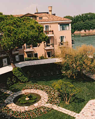 Two paved paths meet at a garden fountain, seen from above in a view that captures the hotel and the adjacent canal.