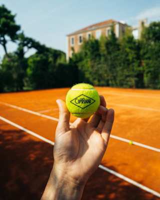 A man's hand holds a Cipriani-branded yellow tennis ball, with the clay-surfaced court and hotel in the background.