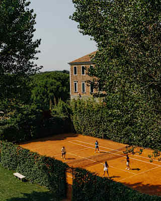 High-angle view over a clay tennis court, bordered by hedgerows and framed by trees, where four players enjoy a game.