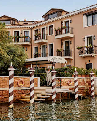 A staff member waves from Hotel Cipriani's awning, seen between the red and white bricole poles in a view from the canal.