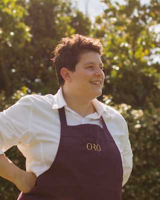 Oro Head Chef Vania Ghedini, with short dark hair, wearing a branded apron, smiles in a portrait taken in the hotel gardens.